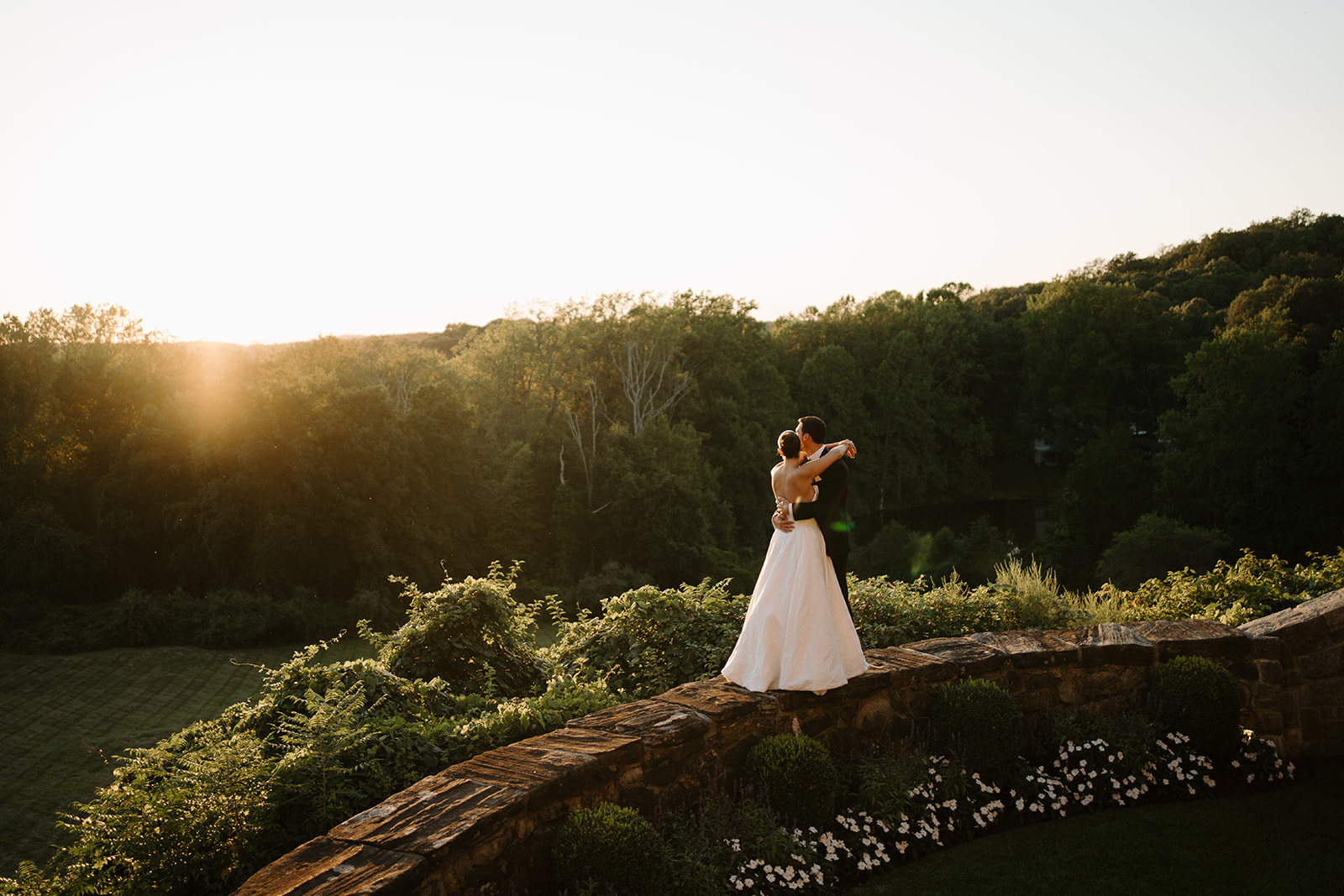 Newlyweds embracing on a stone wall overlooking a lush green valley during a warm, golden sunset.